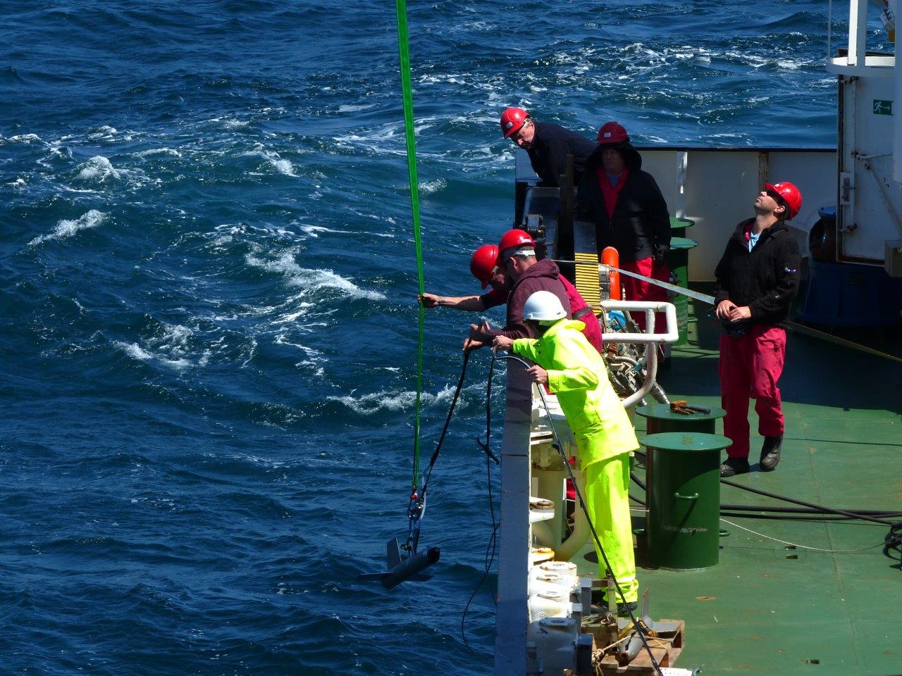 homens-com-capacete-a-bordo-de-um-navio-colocam-aparelho-na-agua-do-mar-com-ajuda-de-um-cabo